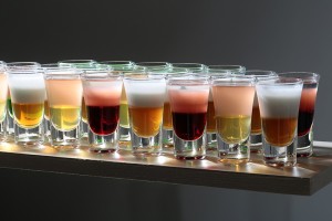 Set of beautiful multicolored red yellow golden and white sweet shots cocktail alcohol drink in glasses standing on bar on grey background, horizontal picture